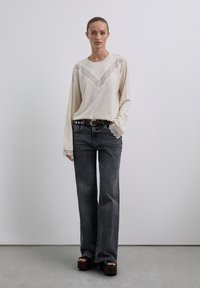 Frau trägt einen cremefarbenen Pullover mit Spitzenbesatz, dunkelgraue weite Jeans, einen genoppten Gürtel und schwarze Plateausandalen und steht vor einer schlichten Wand.