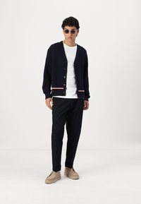 Marineblå ribbestrikket cardigan med hvite og røde stripeakenter i kanten, båret over en hvit t-skjorte, kombinert med mørke bukser og beige sko.