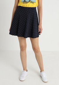 Marinblå kjol med vita prickar, utsvängd design, medellång, tillverkad av mjukt tyg. Matchad med vita sneakers och en gul t-shirt.