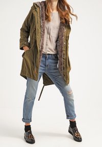 Mujer vistiendo un abrigo parka de piel sintética de color verde oliva, parte superior beige, jeans rasgados con puños, calcetines negros y zapatos oxford de estampado de leopardo, de pie con las manos en los bolsillos.