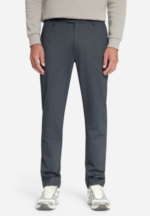 Mann trägt dunkelgraue gerade geschnittene Hose, beige langärmliges Hemd und weiße Sneakers und steht vor weißem Hintergrund.