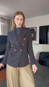 Blazer noir croisé avec des boutons dorés, coupe ajustée, porté sur un col roulé sombre, associé à un pantalon beige plissé large.