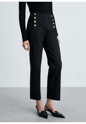 Vrouw draagt korte zwarte broek met decoratieve gouden knopen, gecombineerd met zwarte slingback schoenen met puntige neus.
