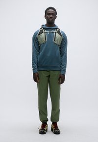 Φόρμα μπλε με εφαρμογή, συνδυασμένη με πράσινο παντελόνι και παπούτσια πεζοπορίας. Ένας ελαφρύς ιμάντας με δύο τσέπες κάθεται στο στήθος. Λευκό φόντο.