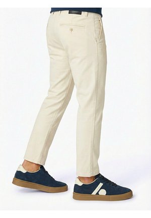 Uomo con pantaloni beige aderenti e sneakers blu navy con dettagli bianchi, fotografato di profilo e da dietro.