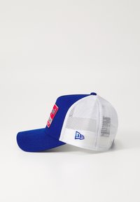 Blå basebollkeps med en böjd skärm, röd patchlogotyp på framsidan, vit nätbakgrund och blå logotypaccent på sidan.