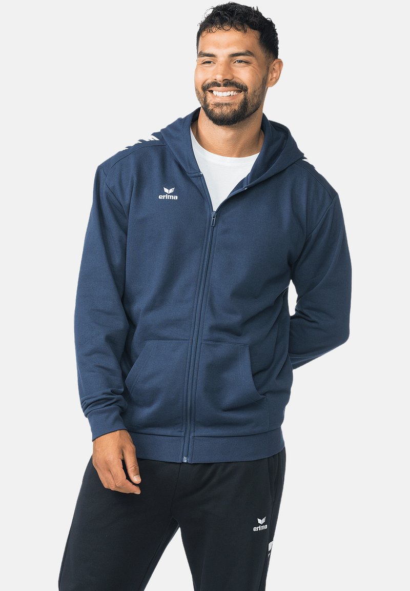 Marineblauwe hoodie met rits, gemaakt van zachte stof, voorzien van een voorzak, verstelbare capuchon en logo op de borst en mouw. Zwarte broek hieronder afgebeeld.