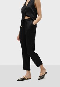 Svart ärmlös top med V-ringning och texturerat mönster, kombinerad med avslappnade svarta byxor och naggad svarta slip-on skor.