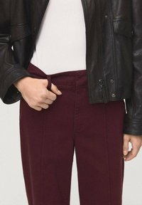 Pantaloni bordeaux con vita aderente, realizzati in tessuto misto cotone, mostrati insieme a una giacca di pelle nera e a una maglietta bianca.