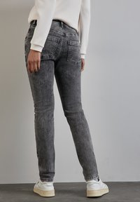 Jean en denim gris au design ajusté, avec des poches arrière et une décoloration subtile, associés à des chaussures blanches sur un sol texturé.
