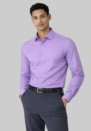 Homme portant une chemise à manches longues lavande boutonnée, rentrée dans un pantalon gris foncé, les mains dans les poches, sur un fond gris.