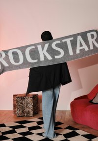 Sciarpa grigia con la scritta "ROCKSTAR" in grandi lettere bianche, appoggiata sulle spalle di una persona. Blazer nero, jeans chiari e uno sgabello a stampa leopardata.