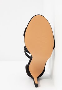 Sandale à talon haut en daim noir montrée par la semelle, avec une semelle beige et un talon fin sur fond blanc.