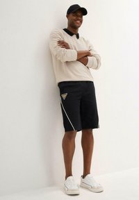 Maglietta beige a maniche lunghe con colletto nero, pantaloni shorts neri con dettagli bianchi e logo, e sneakers bianche. L'individuo è in piedi con le braccia incrociate.
