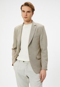 Ljusbeige blazer med texturerad tyg, enkel knappstängning och slagkrage. Bärs över en vit skjorta och ljusgrå byxor.