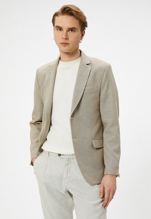 Lys beige blazer med et struktureret stof, lukning med en enkelt knap og klippede slag. Båret over en hvid skjorte og lysegrå bukser.