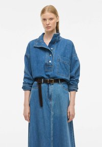Jeans srajca z visokim ovratnikom, sprednjim gumbnim zapiranjem in žepom na prsih, v kombinaciji z dolgo jeans krilo in rjavim pasom, kar ustvarja usklajen modri videz.