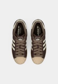 Zapatillas deportivas marrones con una puntera de goma texturizada, tres rayas crema y detalles en beige. Diseño con cordones y plantilla acolchada.