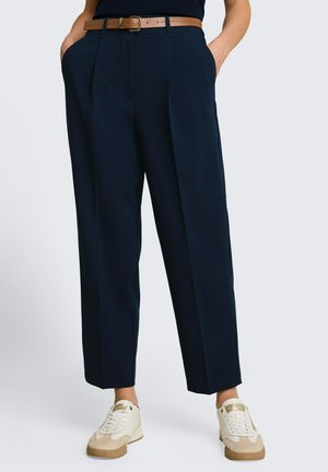 Marineblaue plissierte Hose mit braunem Gürtel, getragen mit beigen und weißen Sneakern, Hände in den Taschen, vor einem schlichten hellen Hintergrund.
