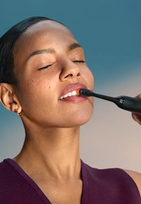 Brosse à dents électrique avec un élégant manche noir, une tête de poils orange et un design ergonomique. Utilisateur appliquant la brosse sur ses dents, sur un fond doux.