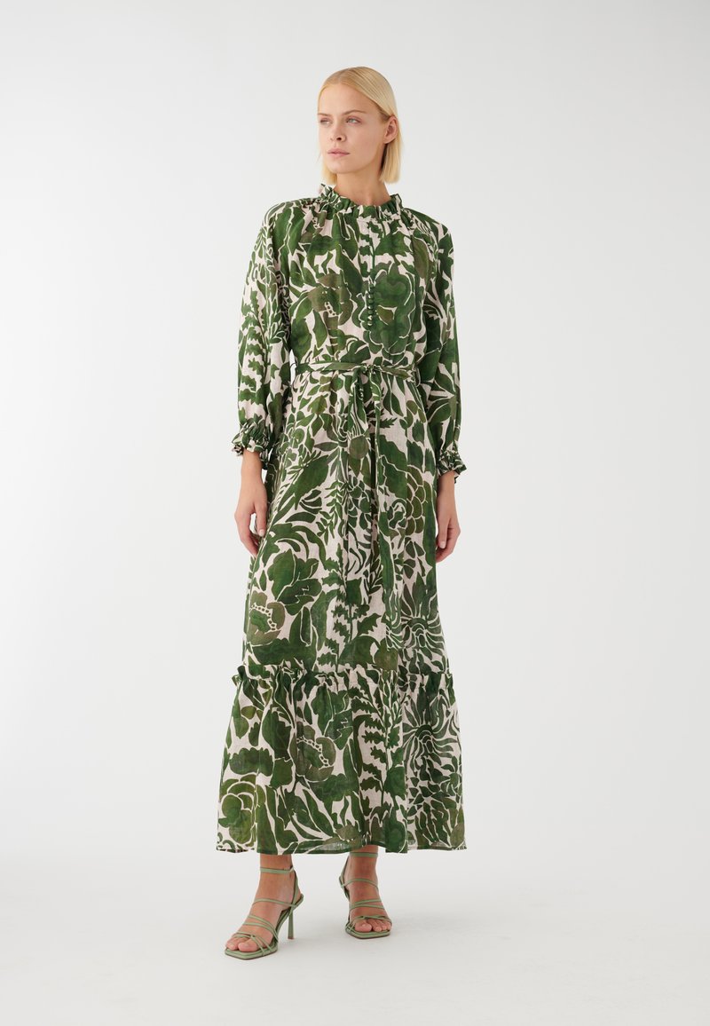 Grünes und cremefarbenes Blumenkleid im Maxiformat mit langen Ärmeln, Rüschenausschnitt und -manschetten, tailliertem Bund und gestuftem Saum; kombiniert mit grünen Riemchensandalen.