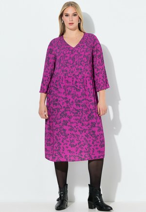 Kobieta stojąca, ubrana w fioletową sukienkę do kolan z czarnym kwiatowym wzorem, czarne rajstopy i czarne botki na białym tle.