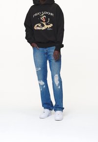 Černá mikina s kapucí s nápisem "PECADOR" a grafikou hada, doplněná světle modrými roztrhanými džínami a bílými teniskami. Minimalistické pozadí.