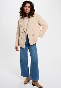 Mujer con cabello rizado, vistiendo una chaqueta beige de doble botonadura, un suéter crema, jeans de pierna amplia azules y botas de tacón color beige, de pie con una mano en la cadera.