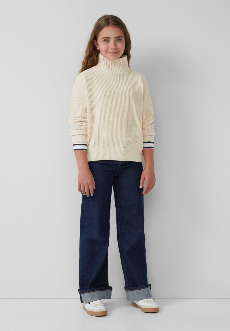 Crème-kleurige geribde coltrui met brede mouwen, gecombineerd met donkere blauwe wijde jeans en witte sneakers met lichtbruine zolen.