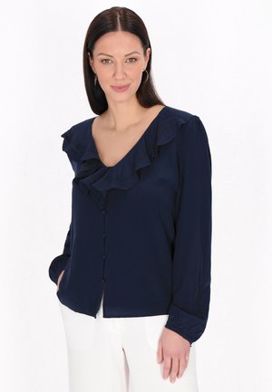 Marinebluse med en volangkrave, lange ærmer og knaplukning, lavet af et glat stof. Modellen står med den ene hånd i lommen.