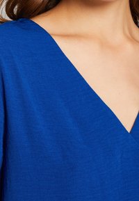 Blauwe linnen kleding met een diepe V-hals en zachte textuur. De stof heeft een lichte glans, minimale naden en schone lijnen.