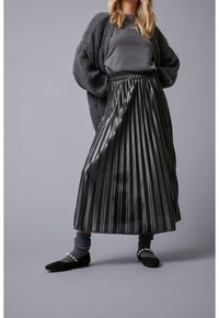 Falda negra con pliegues hecha de un tejido brillante, combinada con un suéter gris y calcetines, y zapatos planos negros con acentos de perlas.