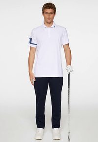 Herrarnas golfpolo, vit med marinblå detaljer på ärmarna, kombinerad med marinblå byxor. Modellen håller i en golfklubba och bär vita skor och handskar.