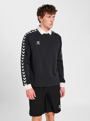 Hombre con sudadera deportiva negra con rayas en forma de cheurón blancas en las mangas, cuello blanco y pantalones cortos negros con logotipos de la marca.