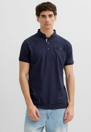 Joven con polo azul marino de manga corta con un pequeño patrón de puntos rojos y vaqueros azul claro, de pie sobre un fondo blanco liso.