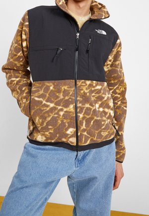 Persoon draagt een fleecejack met een bruin- en beige giraffenpatroon, zwarte borstpanelen, met ritssluitingen uitgeruste zakken en lichtblauwe jeans.