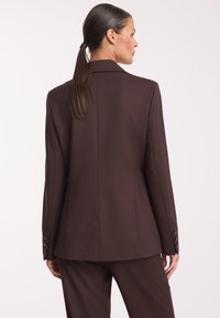 Brązowa marynarka dopasowana o wyraźnej sylwetce, z jednym rozcięciem z tyłu i guziczkami na mankietach. Gładka tekstura materiału.