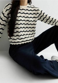 Maglione lavorato a maglia color crema con motivo zigzag blu navy; scollatura rotonda e orlo a coste. Abbinato a pantaloni scuri e calzature bianche.