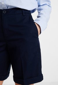 Námořnicky modré bavlněné šortky s klasickým designem, vybavené předním zapínáním na knoflíky, bočními kapsami a ohrnutým lemem. Hladká textura.