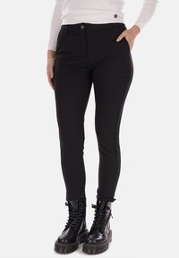 Pantaloni neri aderenti con una texture liscia, dotati di chiusura frontale a bottone, tasche laterali e pannelli laterali a contrasto. Indossati con stivaletti neri con lacci spessi.