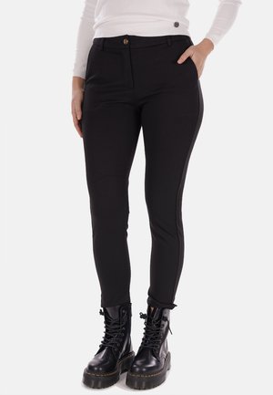 Pantaloni neri aderenti con una texture liscia, dotati di chiusura frontale a bottone, tasche laterali e pannelli laterali a contrasto. Indossati con stivaletti neri con lacci spessi.
