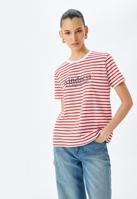 Rød og hvid stribet T-shirt med rund hals, med teksten "venlighed (er en superkraft)" trykt i marineblå. Båret med blå jeans.