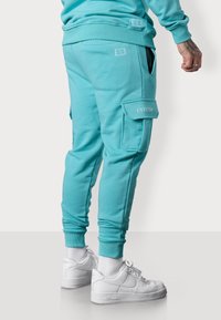 Türkise Sweatpants mit gerippten Bündchen, Cargo-Tasche und Logodetail. Getragen mit weißen Sneakers. Glatte Textur, körpernahe Passform.