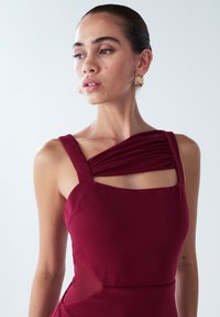 Vyno spalvos suknelė su storomis petnešėlėmis, puošta klostuota petimi ir kvadratinio kirpimo iškirpte. Suknelė turi iškirptes juosmens srityje ir lygios tekstūros audinį.