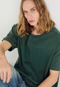 Camiseta de gran tamaño en verde oscuro, hecha de algodón, con cuello redondo y mangas cortas. Textura suave y lisa con un corte relajado.