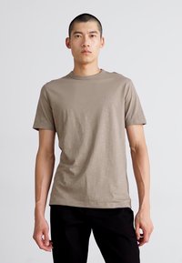 Tricou ajustat, maro, cu mâneci scurte, guler rotund și textură subtilă. Are un mic logo pe piept. Asortat cu pantaloni negri.