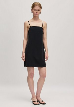 Zwarte slipdress met dunne bandjes, rechte pasvorm, knielang, gladde textuur, subtiele glans, met kleine hardware accenten op de bandjes.