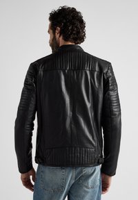 Zwarte leren jas met een gewatteerd ontwerp op de schouders en rug. Heeft een getailleerde pasvorm en een gladde textuur. Gecombineerd met lichtblauwe jeans.