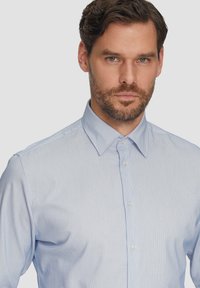 Svijetloplena košulja s dugim rukavima, s malo vidljivom teksturom, uskim ovratnikom i prozirnim dugmadima, s krojem koji naglašava figure.