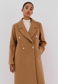 Cappotto doppiopetto in misto lana cammello, con ampia revers e due tasche frontali, oltre a bottoni in tonalità argento. Texture morbida.